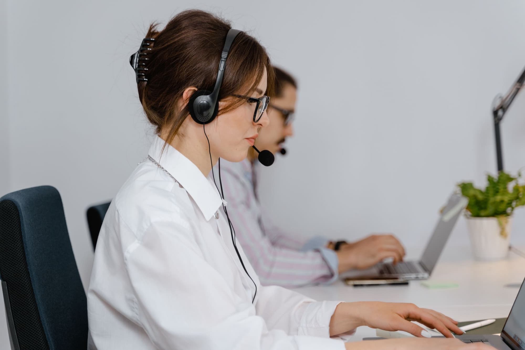 Contact-centre agent in profile wearing a headset, typing at a laptop beside a colleague