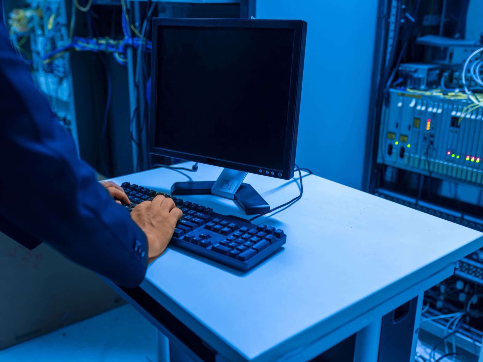 Technician at a monitoring console beside racks of network equipment in a blue-lit server room
